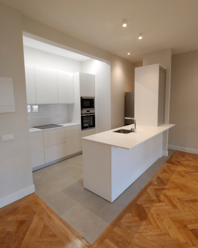 Imagen de una cocina reformada por MAM Experience Building, destacando una isla central con fregadero, electrodomésticos integrados, y un elegante suelo de madera que se extiende hacia el área de la sala, creando un espacio abierto y funcional.